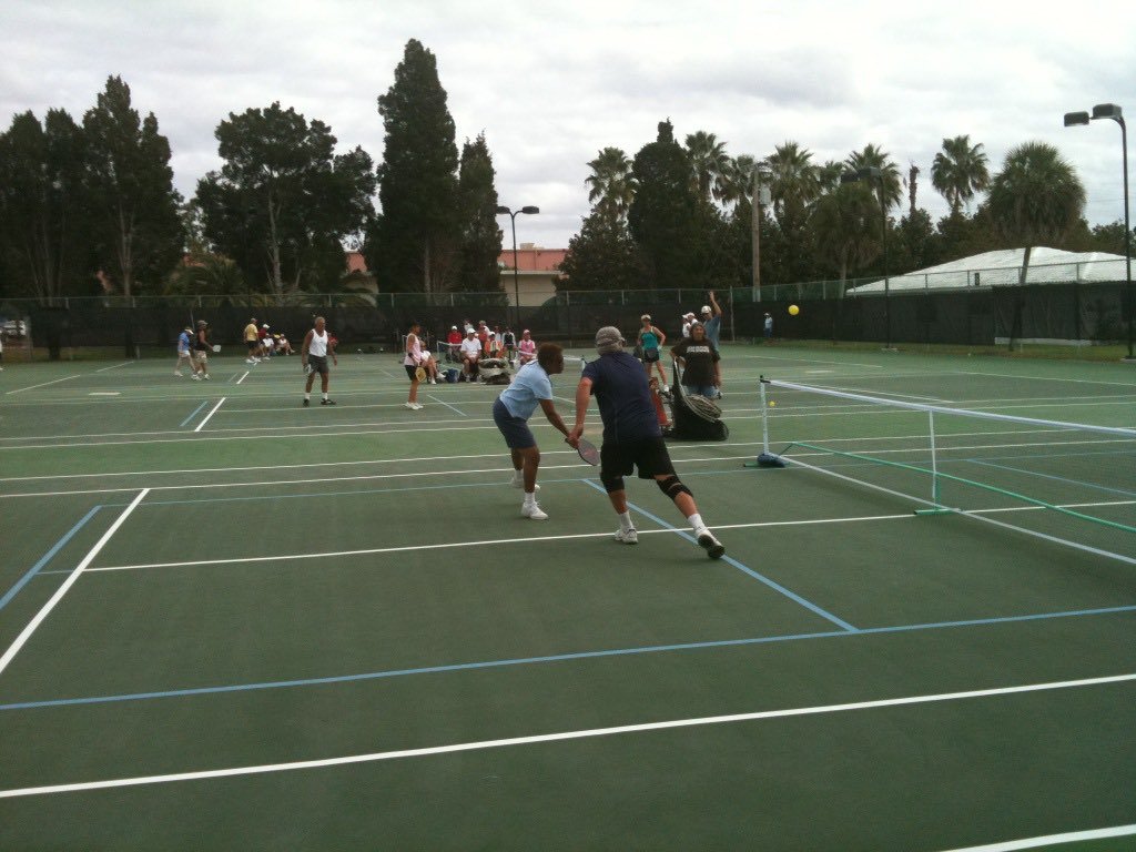 doubles rally on an outdoor pickleball court, all four players at the kitchen line